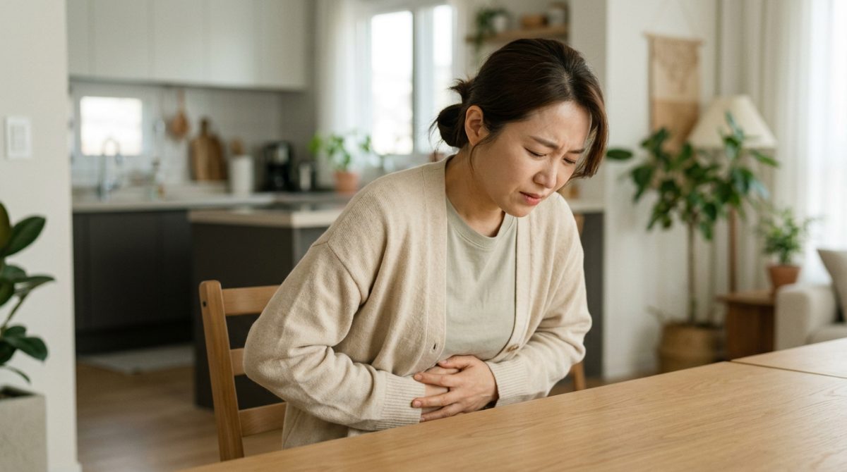 깔끔하고 현대적인 집안의 빈 테이블 앞에서 소화불량과 복통으로 인해 두 손으로 배를 움켜잡고 힘들어하는 30대 여성의 모습